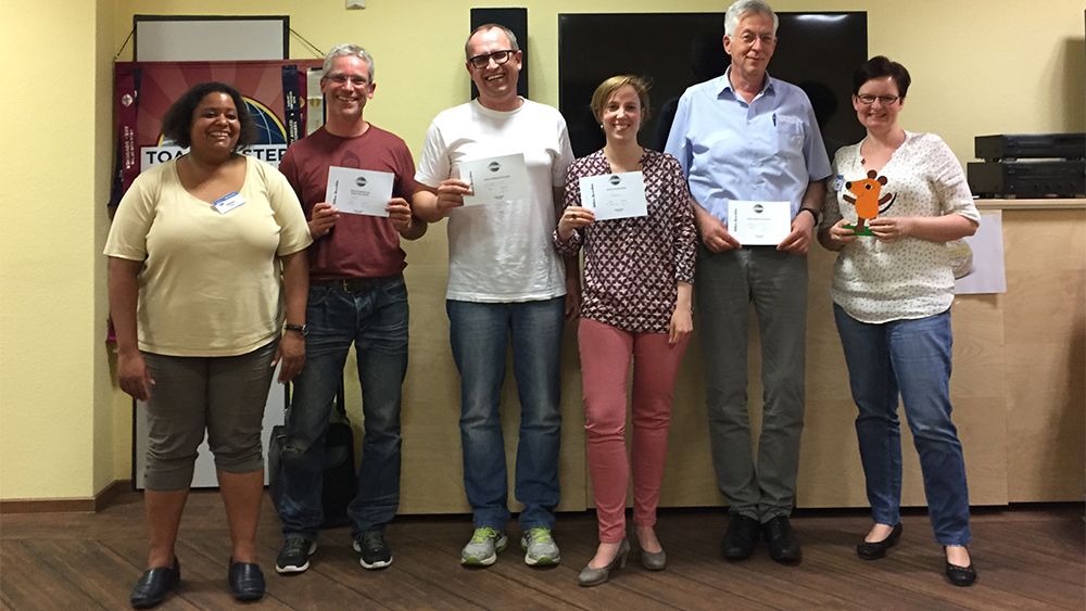 Sechs lächelnde Mitglieder der Kölner Sprechbar stehen nebeneinander vor dem Toastmasters-Banner und halten ihre Urkunden in den Händen.
