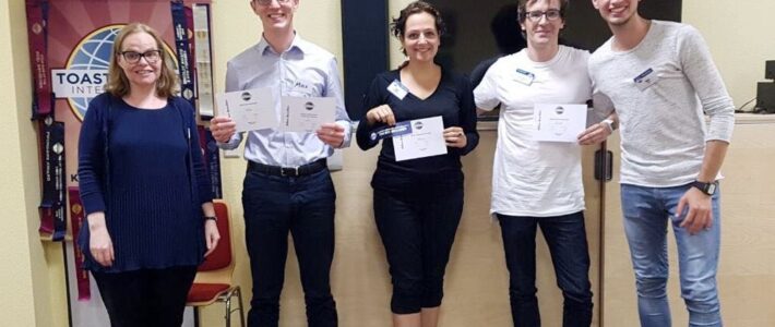 Fünf lächelnde Mitglieder der Kölner Sprechbar stehen vor dem Toastmasters-Banner und halten ihre Urkunden in den Händen.