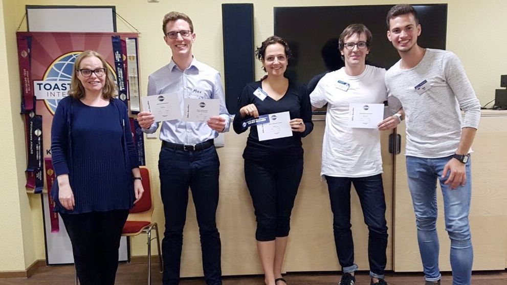 Fünf lächelnde Mitglieder der Kölner Sprechbar stehen vor dem Toastmasters-Banner und halten ihre Urkunden in den Händen.