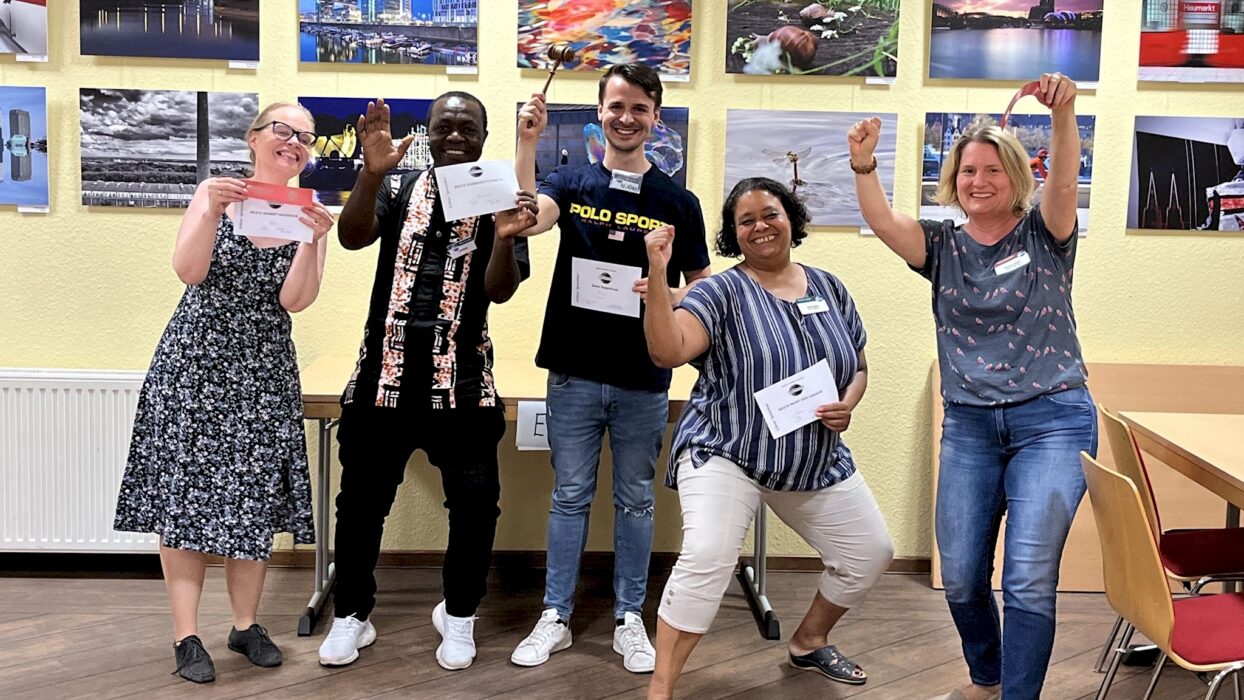 Fünf jubelnde Toastmasters-Mitglieder strecken die Fäuste in die Luft und halten ihre Auszeichnungsurkunden, im Hintergrund eine Fotoausstellung.