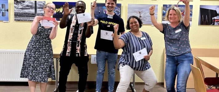 Ehrenamt – Was uns zusammenhält! Fünf jubelnde Toastmasters-Mitglieder strecken die Fäuste in die Luft und halten ihre Auszeichnungsurkunden, im Hintergrund eine Fotoausstellung.