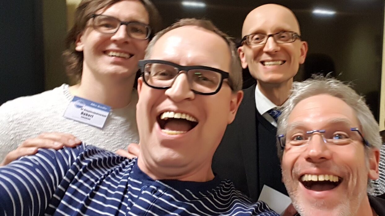 Vier lächelnde Mitglieder der Kölner Sprechbar posieren eng zusammen für ein Selfie in geselliger Runde.