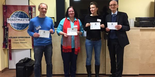 Vier lächelnde Personen stehen vor einem Toastmasters-Banner und halten ihre Urkunden in den Händen.