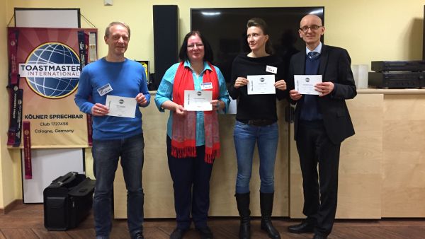 Vier lächelnde Personen stehen vor einem Toastmasters-Banner und halten ihre Urkunden in den Händen.