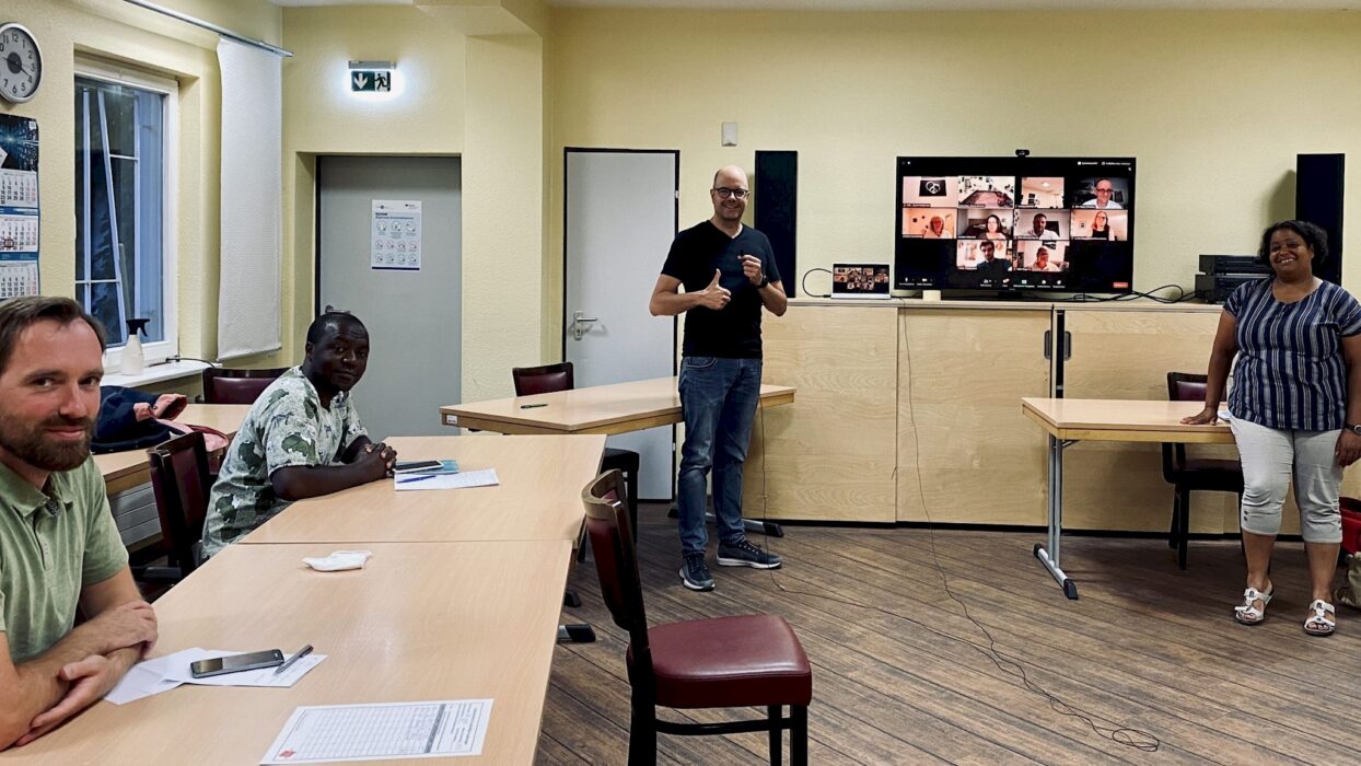 Toastmasters-Mitglieder sitzen im Meetingraum der Kölner Sprechbar, während auf einem Bildschirm im Hintergrund weitere Mitglieder per Zoom zugeschaltet sind.