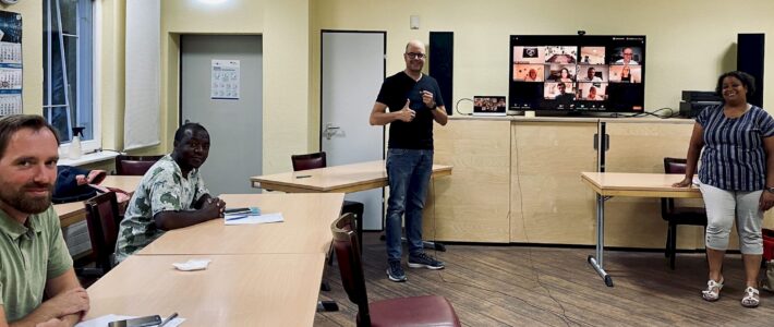 Toastmasters-Mitglieder sitzen im Meetingraum der Kölner Sprechbar, während auf einem Bildschirm im Hintergrund weitere Mitglieder per Zoom zugeschaltet sind.