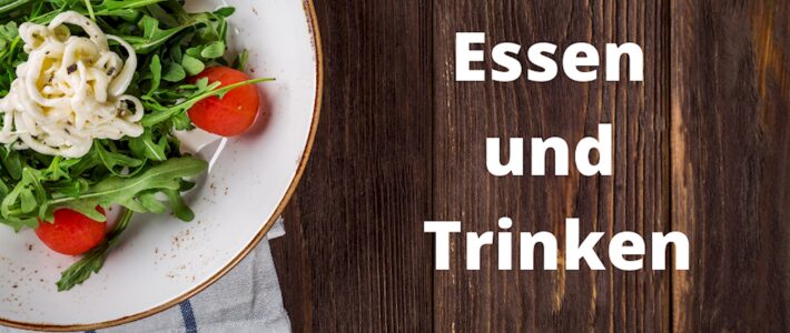Teller mit Salat und Besteck auf einem Holztisch neben dem Schriftzug Essen und Trinken.