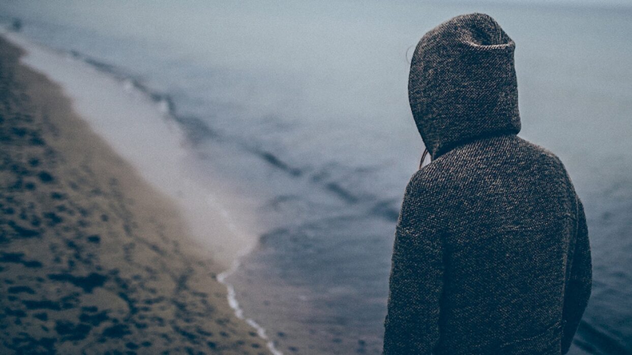 Person im Kapuzenmantel steht mit dem Rücken zur Kamera an einem Strand.