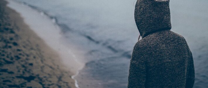 Auch das ist ein Teil von mir Person im Kapuzenmantel steht mit dem Rücken zur Kamera an einem Strand.