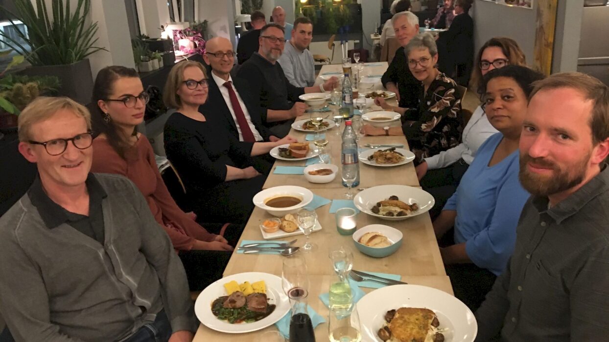 Eine Gruppe von etwa 13 Personen sitzt gemeinsam an einer langen Restauranttafel mit Vorspeisen und Getränken zur 10-Jahres-Feier der Kölner Sprechbar.