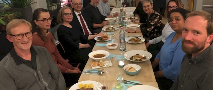 Eine Gruppe von etwa 13 Personen sitzt gemeinsam an einer langen Restauranttafel mit Vorspeisen und Getränken zur 10-Jahres-Feier der Kölner Sprechbar.