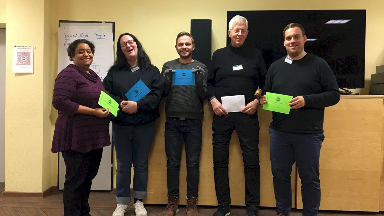 Fünf lächelnde Toastmasters-Mitglieder mit bunten Auszeichnungskarten stehen nebeneinander vor einem Flipchart.