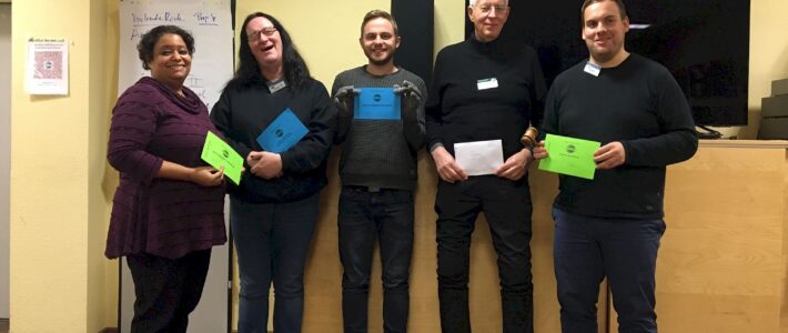 Fünf lächelnde Toastmasters-Mitglieder mit bunten Auszeichnungskarten stehen nebeneinander vor einem Flipchart.