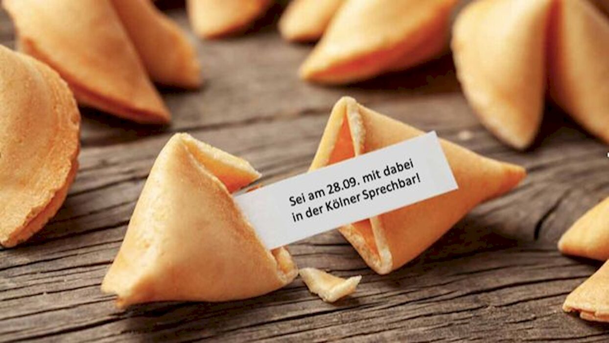 Aufgebrochene Glückskekse auf einem Holztisch, einer davon mit einem Zettel „Sei am 28.09. mit dabei in der Kölner Sprechbar!“.
