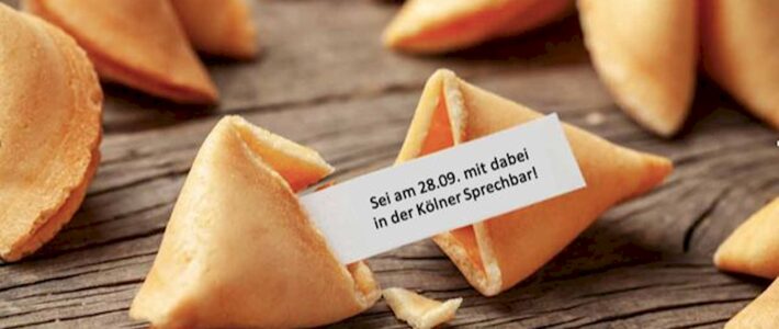 Aufgebrochene Glückskekse auf einem Holztisch, einer davon mit einem Zettel „Sei am 28.09. mit dabei in der Kölner Sprechbar!“.