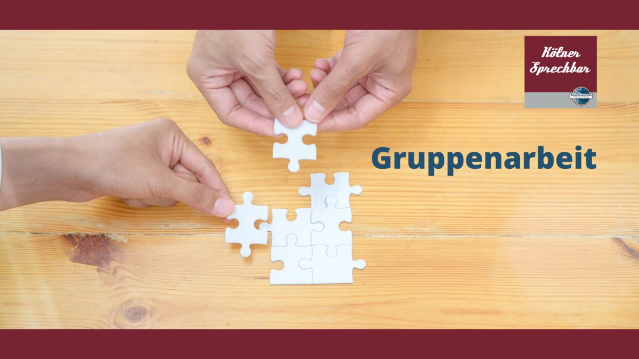 Zwei Hände setzen gemeinsam Puzzleteile auf einem Holztisch zusammen, daneben der Schriftzug „Gruppenarbeit“ und das Logo der Kölner Sprechbar.