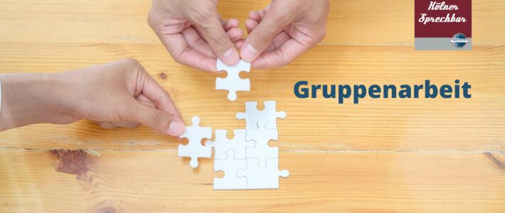 Gruppenarbeit Zwei Hände setzen gemeinsam Puzzleteile auf einem Holztisch zusammen, daneben der Schriftzug „Gruppenarbeit“ und das Logo der Kölner Sprechbar.