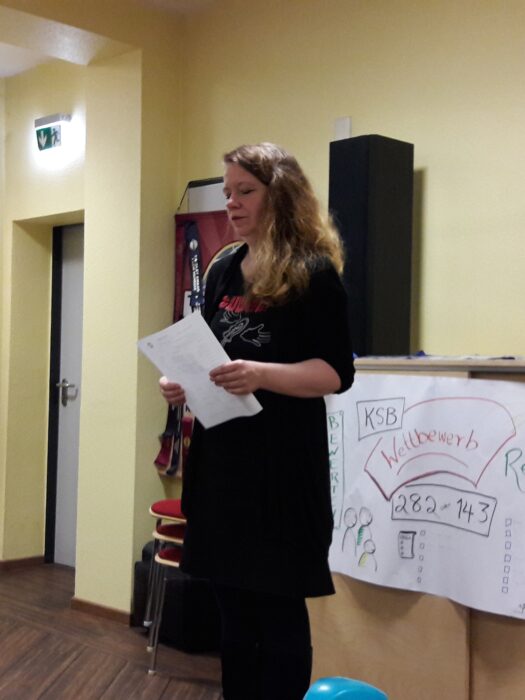 Renate moderiert den Abend und liest von einem Blatt Papier ab, im Hintergrund eine Flipchart mit dem KSB-Wettbewerbsstand.