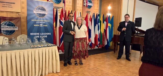 Andrea Sauerzweig schüttelt bei der Distrikt-Konferenz die Hand des Toastmasters-International-Präsidenten Balraj Arunasalam, im Hintergrund die Flaggen der teilnehmenden Länder.