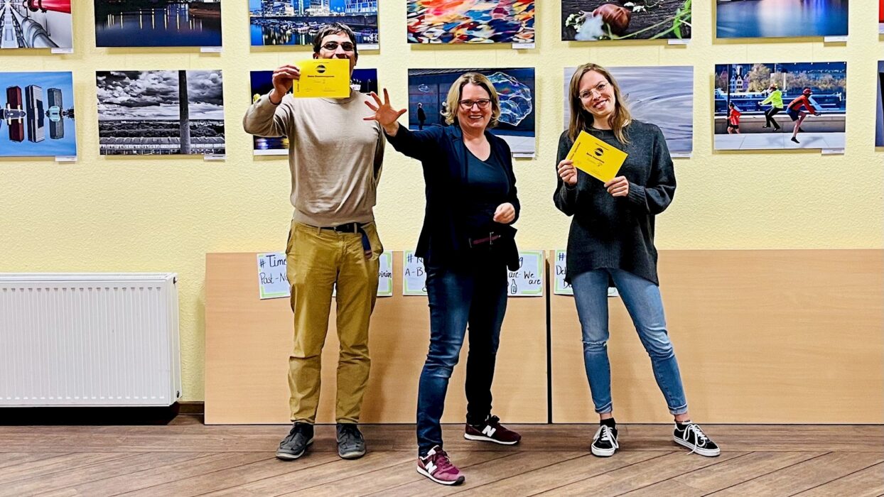 Drei lächelnde Toastmasters-Mitglieder vor einer Fotoausstellung, zwei halten Auszeichnungsurkunden, im Hintergrund Flipchart-Poster mit Stegreifreden-Tricks.