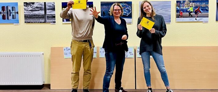 Drei lächelnde Toastmasters-Mitglieder vor einer Fotoausstellung, zwei halten Auszeichnungsurkunden, im Hintergrund Flipchart-Poster mit Stegreifreden-Tricks.