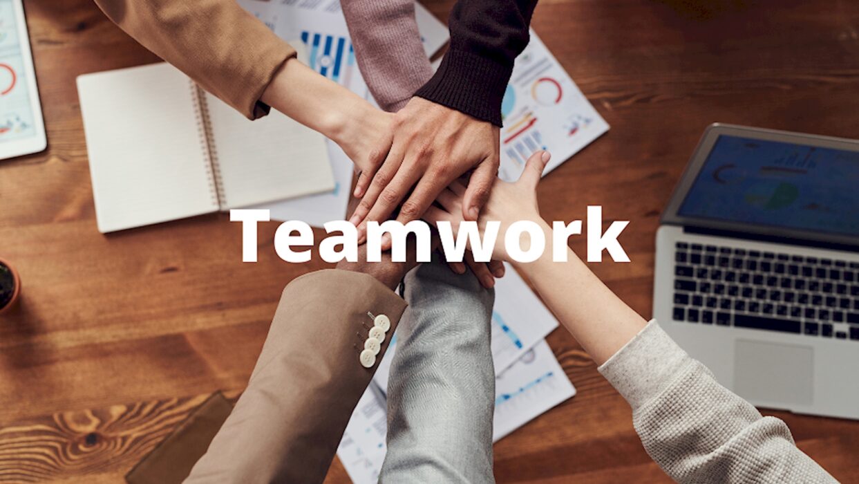 Mehrere Hände legen sich von oben gesehen über einem Schreibtisch mit Diagrammen und Laptop übereinander, dazu der Schriftzug „Teamwork“.