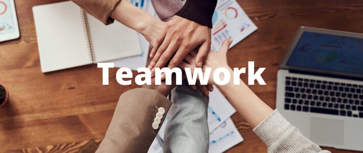 Im Team arbeiten Mehrere Hände legen sich von oben gesehen über einem Schreibtisch mit Diagrammen und Laptop übereinander, dazu der Schriftzug „Teamwork“.