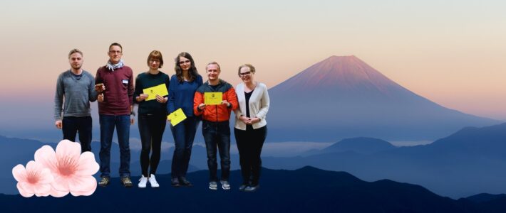 Sechs Toastmasters-Mitglieder mit gelben Auszeichnungskarten stehen vor einer Fotomontage mit dem Fuji und Kirschblüten.
