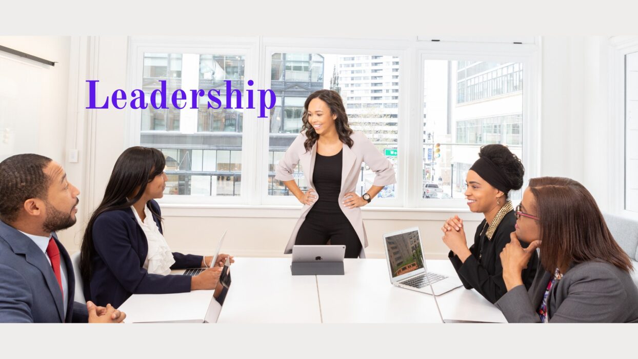 Fünf Personen sitzen an einem Konferenztisch, eine Frau steht mit den Händen in die Hüften gestützt und leitet das Meeting; daneben der Schriftzug „Leadership“.