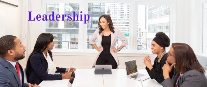 Leadership Fünf Personen sitzen an einem Konferenztisch, eine Frau steht mit den Händen in die Hüften gestützt und leitet das Meeting; daneben der Schriftzug „Leadership“.