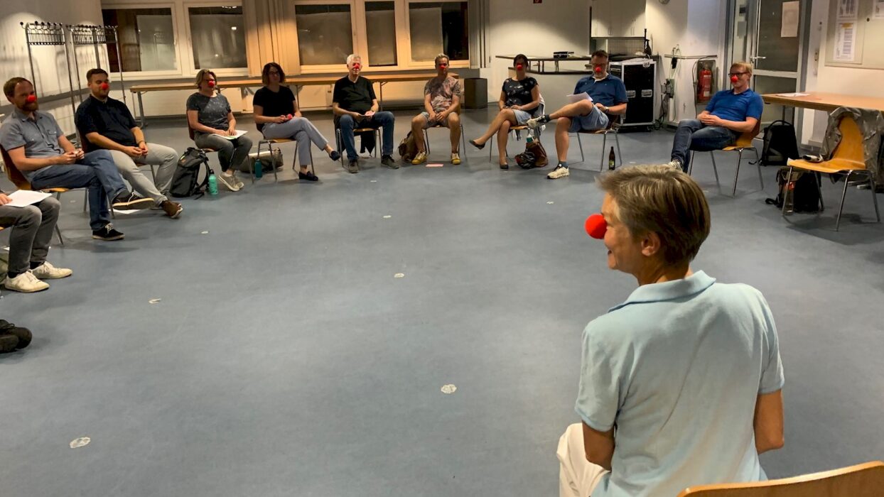Eine Gruppe von Toastmasters-Mitgliedern sitzt im Kreis, alle tragen rote Clownsnasen, während eine Trainerin im Vordergrund den Workshop leitet.
