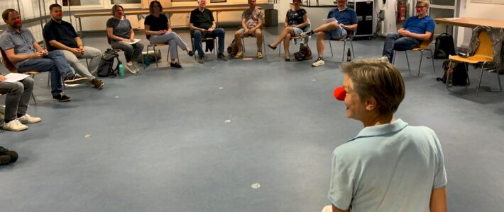 Eine Gruppe von Toastmasters-Mitgliedern sitzt im Kreis, alle tragen rote Clownsnasen, während eine Trainerin im Vordergrund den Workshop leitet.