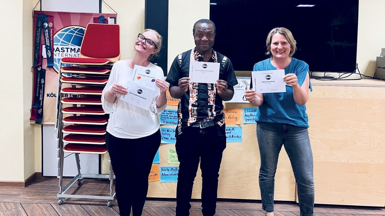 Drei lächelnde Toastmasters-Mitglieder halten ihre Auszeichnungsurkunden, im Hintergrund Klebezettel mit Lieblingsbuchtiteln.