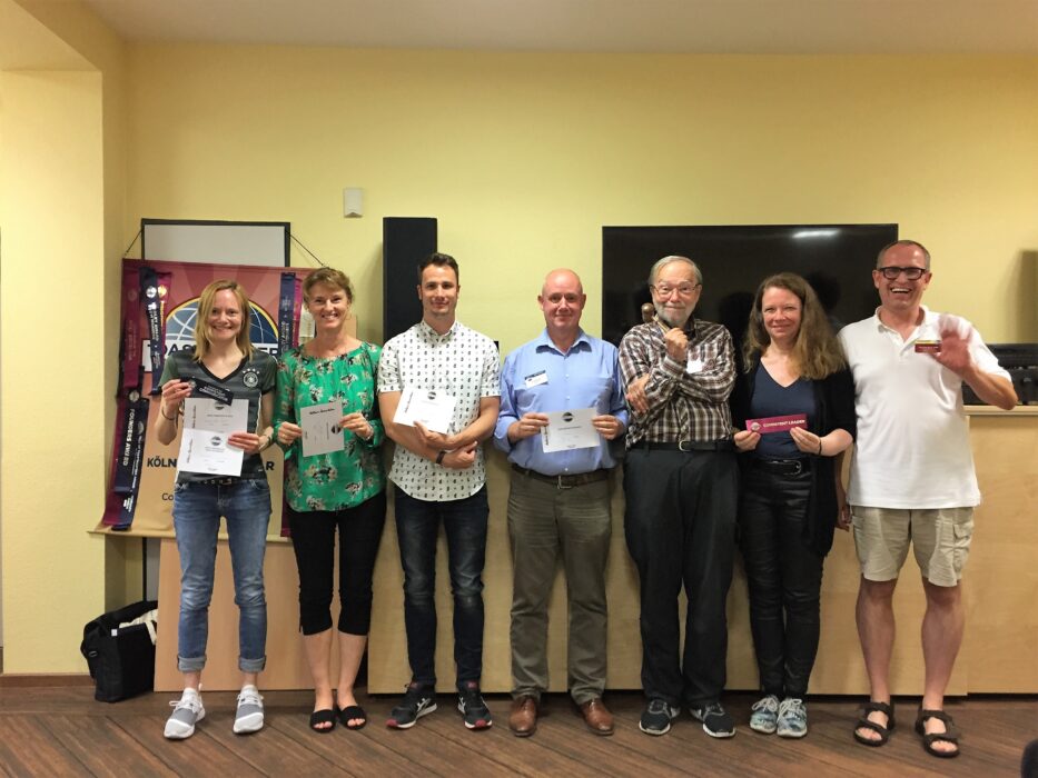 Sieben Mitglieder der Kölner Sprechbar stehen lächelnd nebeneinander vor dem Toastmasters-Banner und halten ihre Urkunden und Abzeichen in den Händen.
