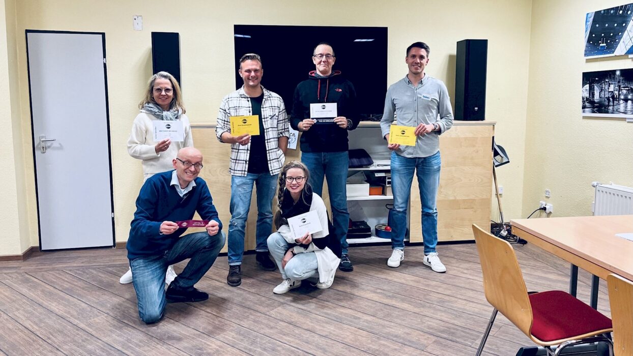 Sechs lächelnde Toastmasters-Mitglieder mit ihren Auszeichnungsurkunden und -bändern, drei stehend im Hintergrund, drei kniend im Vordergrund.