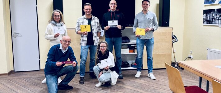 Sechs lächelnde Toastmasters-Mitglieder mit ihren Auszeichnungsurkunden und -bändern, drei stehend im Hintergrund, drei kniend im Vordergrund.