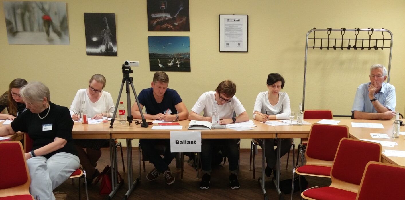 Bewerterinnen und Bewerter sitzen konzentriert schreibend an einem Tisch mit dem Schild "Ballast", dem Wort des Abends.