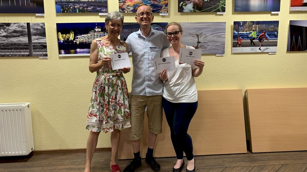 Drei lächelnde Toastmasters-Mitglieder stehen nebeneinander vor einer Fotoausstellung an der Wand und halten ihre Auszeichnungsurkunden in den Händen.
