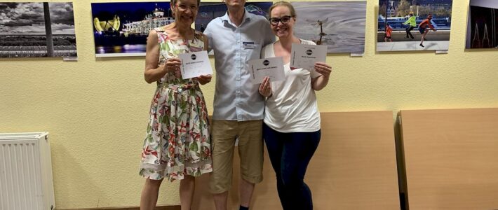 Money makes the world go around… Drei lächelnde Toastmasters-Mitglieder stehen nebeneinander vor einer Fotoausstellung an der Wand und halten ihre Auszeichnungsurkunden in den Händen.