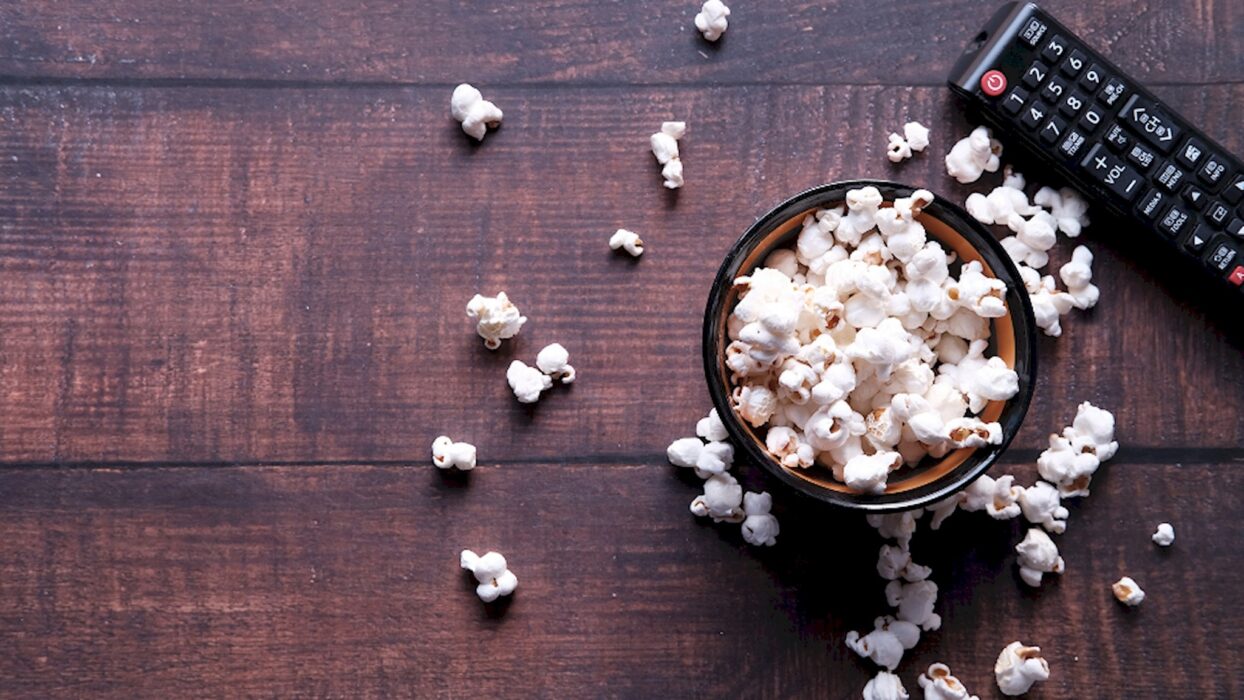 Schale mit Popcorn, verstreutem Popcorn und Fernbedienung auf einem dunklen Holztisch.