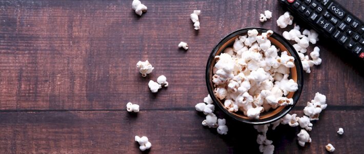 Motion Pictures Schale mit Popcorn, verstreutem Popcorn und Fernbedienung auf einem dunklen Holztisch.