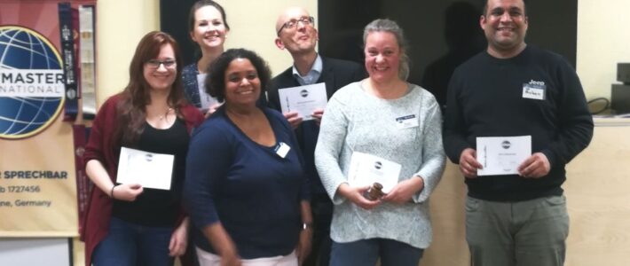 Sechs lächelnde Mitglieder der Kölner Sprechbar stehen vor dem Toastmasters-Banner und halten ihre Urkunden in den Händen.