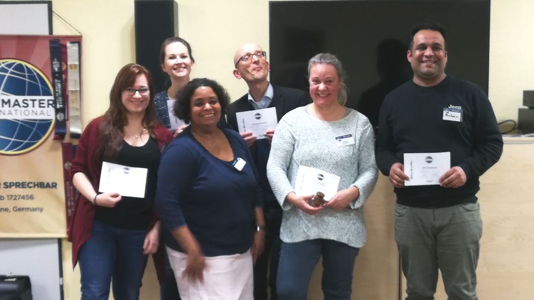 Sechs lächelnde Mitglieder der Kölner Sprechbar stehen vor dem Toastmasters-Banner und halten ihre Urkunden in den Händen.