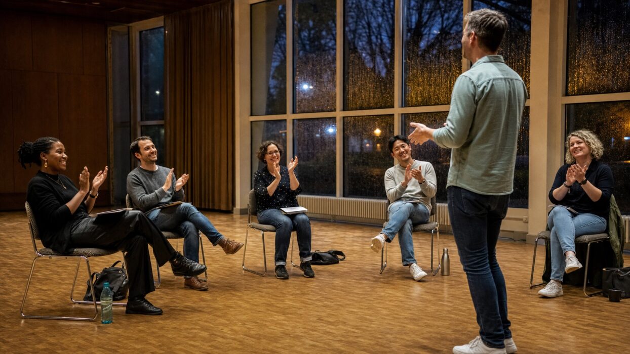 Sechs Erwachsene applaudieren einem Redner beim ersten Präsenztreffen in einem regennassen Saal des Bürgerzentrums.