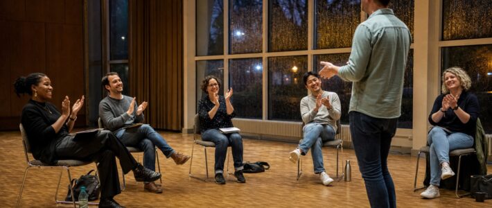 Erstes Präsenztreffen: Freiheit im Bürgerzentrum Sechs Erwachsene applaudieren einem Redner beim ersten Präsenztreffen in einem regennassen Saal des Bürgerzentrums.