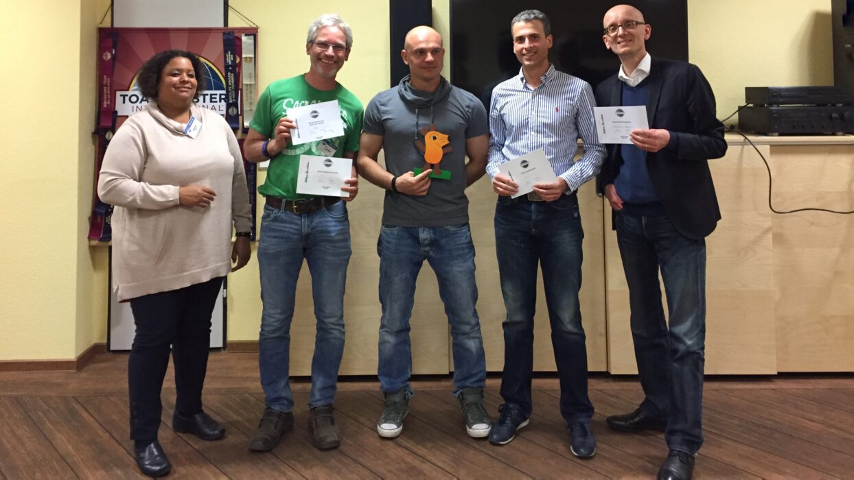 Fünf Mitglieder der Kölner Sprechbar stehen lächelnd nebeneinander vor dem Toastmasters-Banner und halten ihre Urkunden in den Händen.