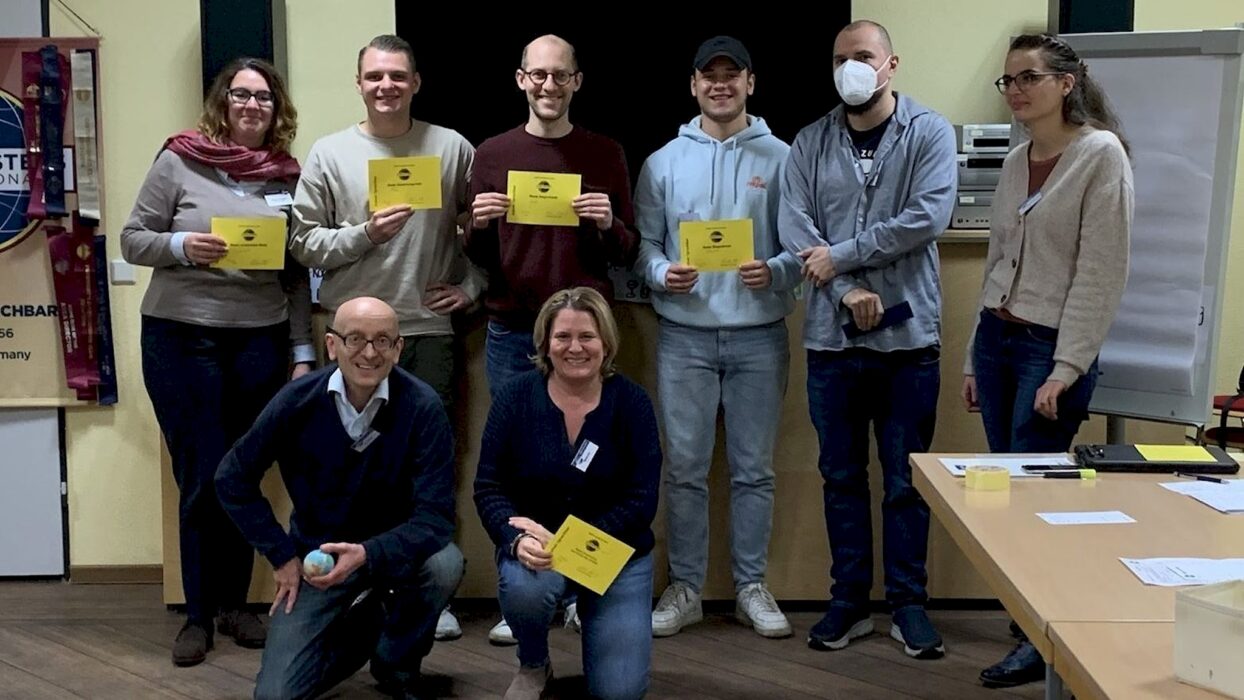 Acht lächelnde Toastmasters-Mitglieder, mehrere mit gelben Auszeichnungsurkunden, posieren gemeinsam vor Vereinsbannern.