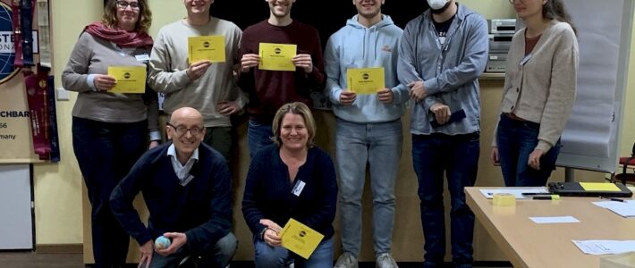 Acht lächelnde Toastmasters-Mitglieder, mehrere mit gelben Auszeichnungsurkunden, posieren gemeinsam vor Vereinsbannern.