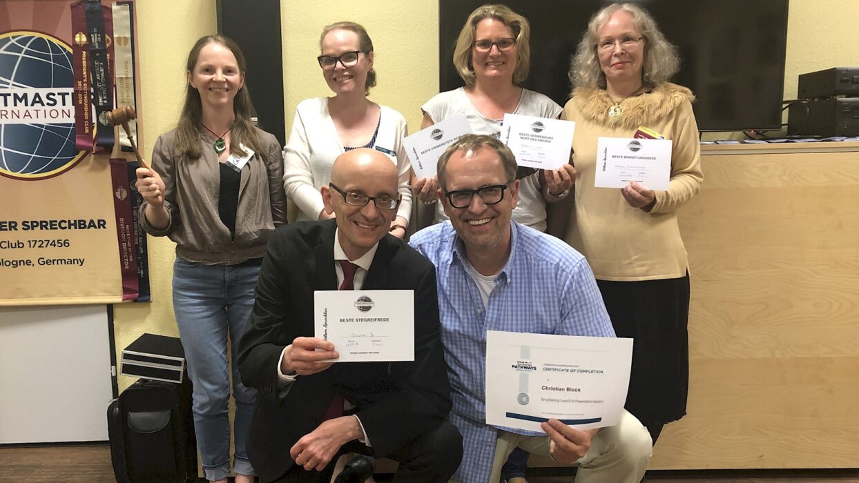 Sechs Mitglieder der Kölner Sprechbar posieren vor einem Toastmasters-Banner mit Urkunden und einem Hammer, zwei Personen knien vorne mit Zertifikaten in der Hand.