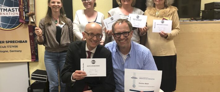 Reflektieren Sechs Mitglieder der Kölner Sprechbar posieren vor einem Toastmasters-Banner mit Urkunden und einem Hammer, zwei Personen knien vorne mit Zertifikaten in der Hand.